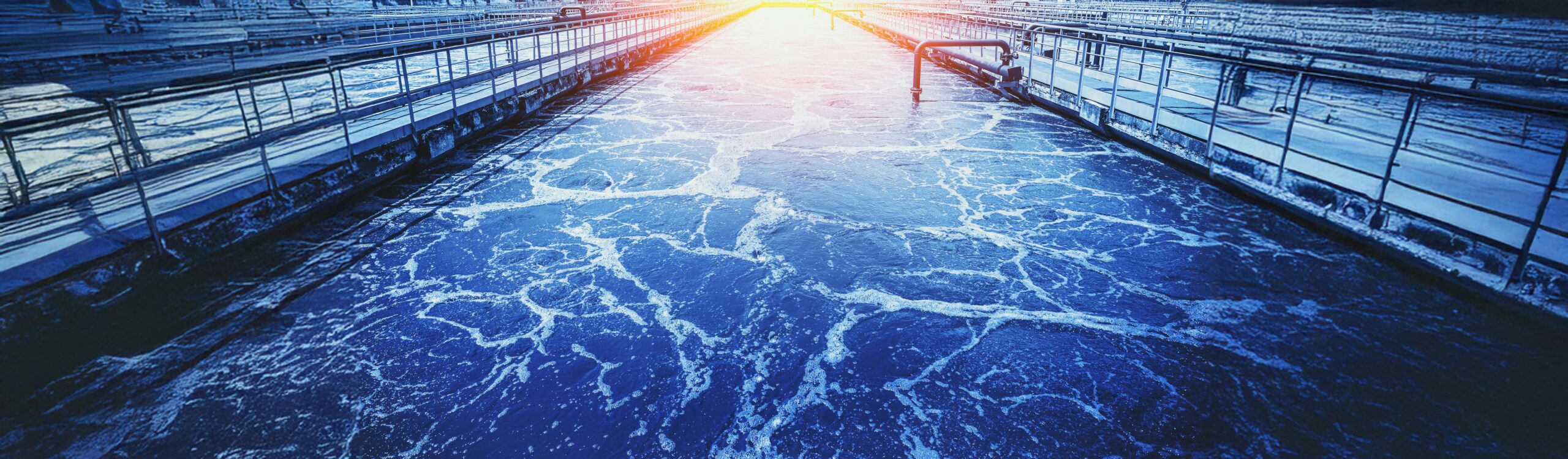 L’image représente un bassin de décantation ou d’aération situé dans une station d’épuration des eaux usées, vue en perspective centrale. On y voit un canal rectangulaire rempli d’eau sombre et agitée, parcourue de nombreuses traces de mousse blanche en surface, formant des motifs organiques. Ces bulles et tourbillons indiquent une activité biologique intense, typique des étapes de traitement aérobie. De chaque côté du bassin, de longues passerelles métalliques équipées de garde-corps permettent la circulation du personnel pour la surveillance et la maintenance. Leurs structures sont régulières, parallèles, et délimitent visuellement le canal principal. Le regard est guidé vers le point de fuite au centre, où une lumière vive et diffuse simule le lever ou coucher du soleil, accentuant la symétrie de la scène. L’ensemble du décor est baigné dans une dominante bleu froid, renforçant l’impression de technicité, de contrôle et de processus industriel. À droite, un tuyau vertical émerge du bord du bassin, équipé d’un robinet ou d’une vanne de régulation. L’ensemble donne à voir une infrastructure d’ingénierie hydraulique avancée, propre et structurée, dans le cadre d’un traitement moderne des eaux usées, probablement en milieu urbain ou industriel.